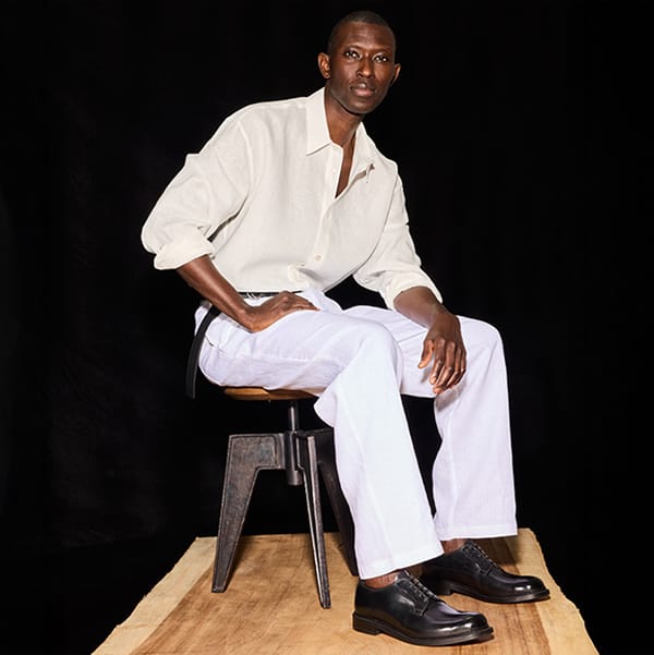 Allen Edmonds Brand man posing on a stool wearing white clothing and a dark background wearing brand dress shoes